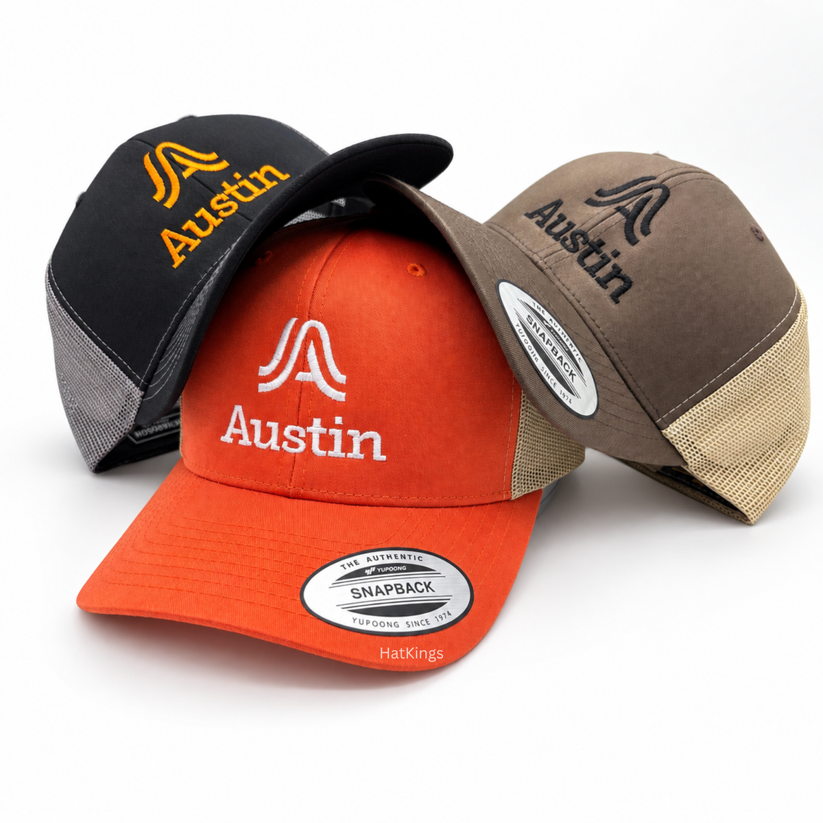 Three yupoong trucker hats in black, orange, and brown with embroidered Austin logos.